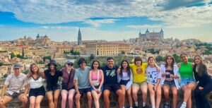 Thirteen students sit on a stone ledge with their arms around each other. In the background is a large city with historic architecture. The sky is bright blue with large white clouds.