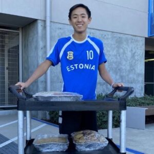 A young man standing behind a cart that has several large trays of food on it